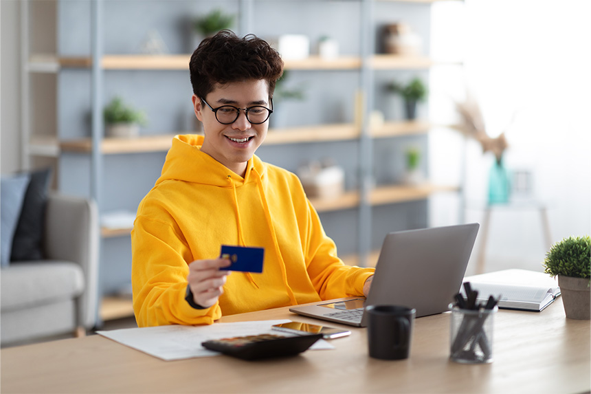 A young man smiles at his credit card as he faces an open laptop.