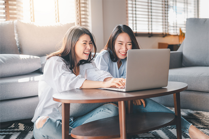 Two young girls smile as they browse on a laptop while sitting on their living room floor.