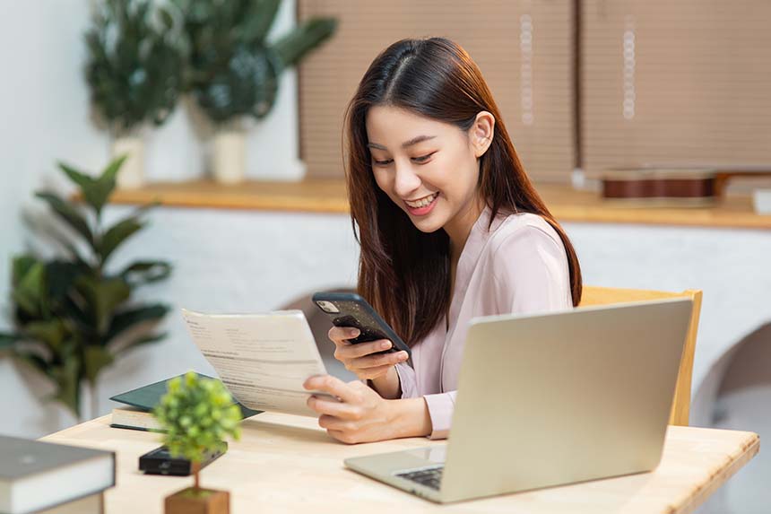 A woman smiling while holding a smartphone and reading a document at a desk with a laptop.