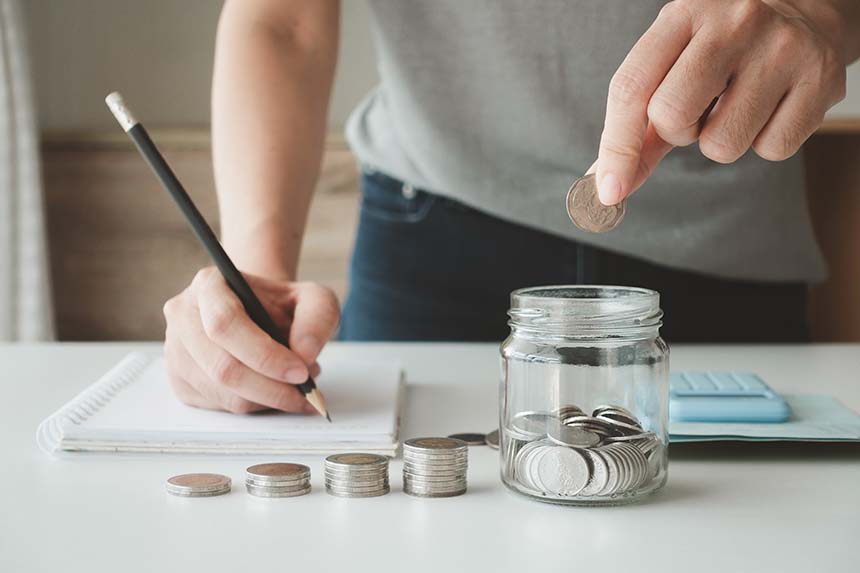 A jar filled with silver coins on a table, with a person holding a paper and a smartphone in the background.