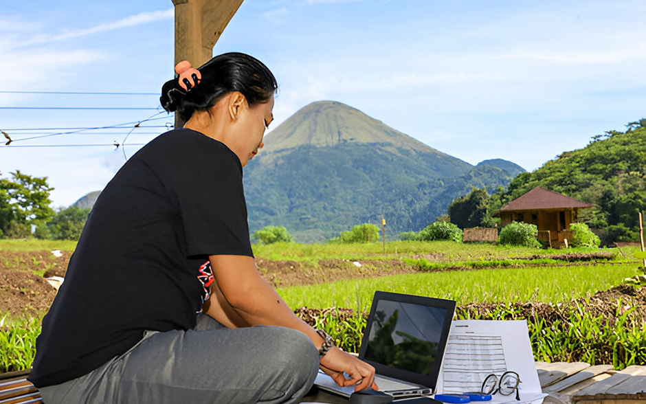 An Asian woman works outdoors with a laptop, set against the backdrop of rice fields and mountains.