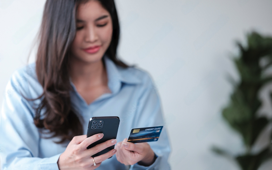 A close-up of an Asian woman using her smartphone and credit card to make an online payment.
