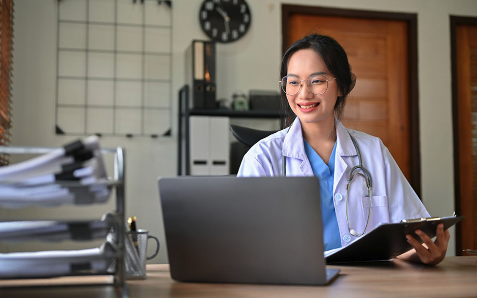 A professional Asian female doctor works in her office, using a laptop to check medical records.
