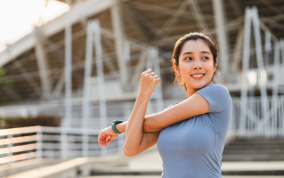 A young Asian woman in sportswear stretches in front of a stadium, warming up before her morning workout with a confident and focused expression.