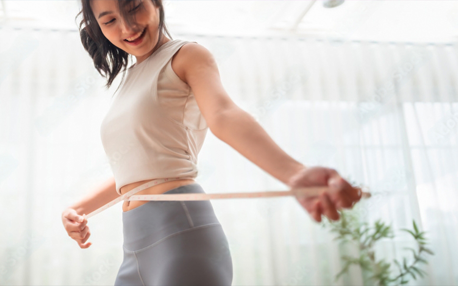 Woman smiling while measuring her waist with a measuring tape indoors.