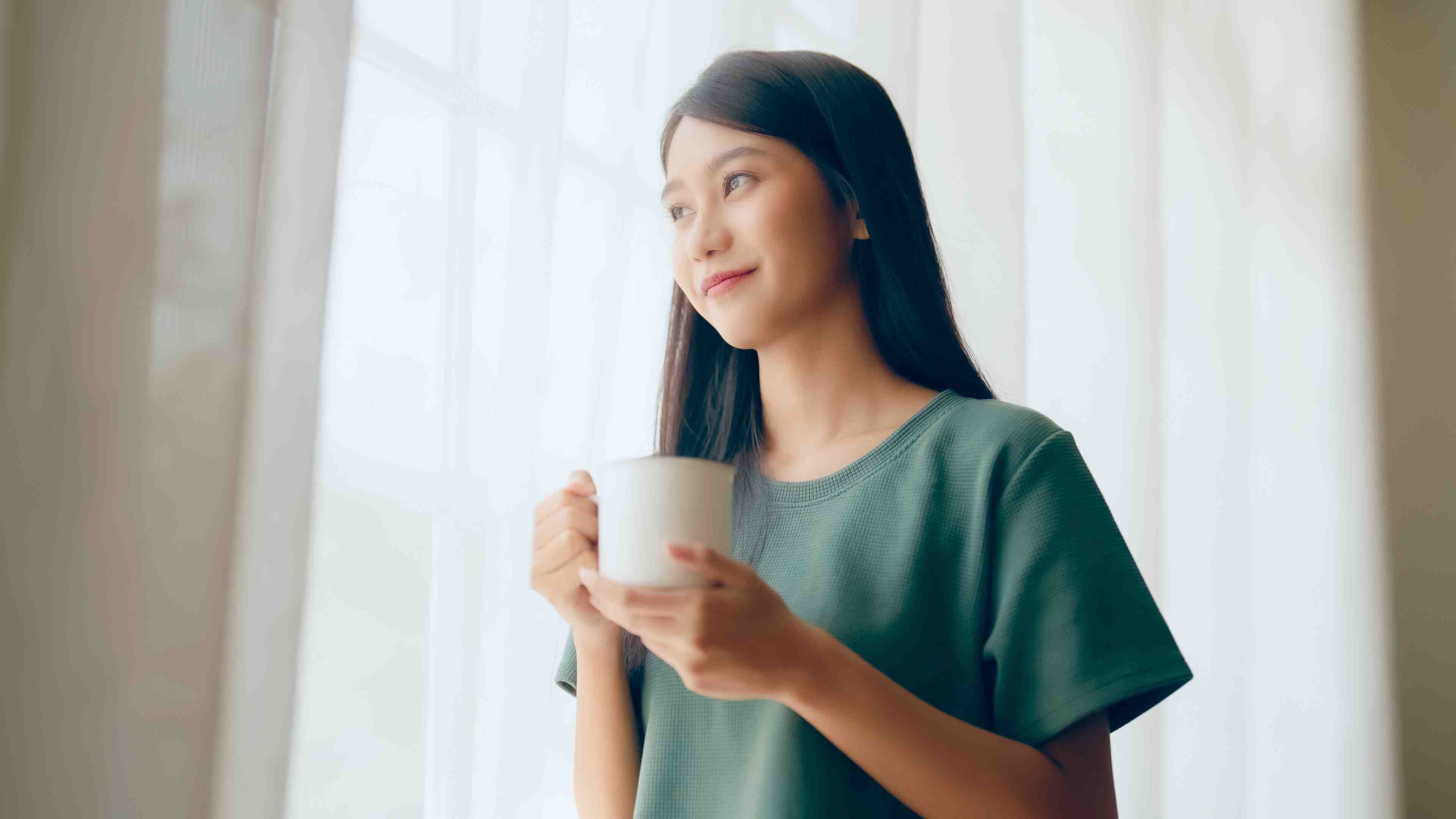 A woman looking out the window while drinking from a cup.