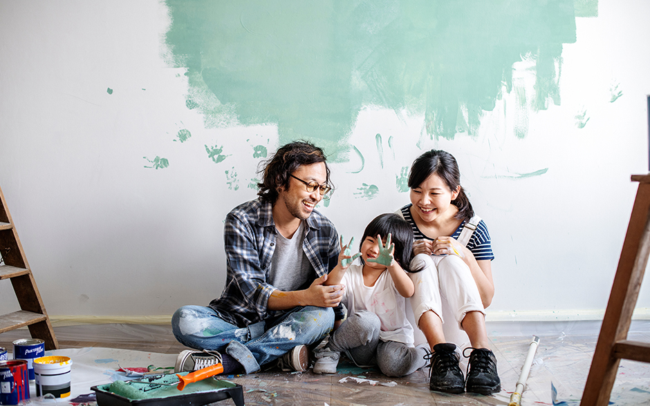 A family doing home renovations, sitting on the floor surrounded by paint supplies, with mom and dad laughing as their child gestures with paint-covered hands.