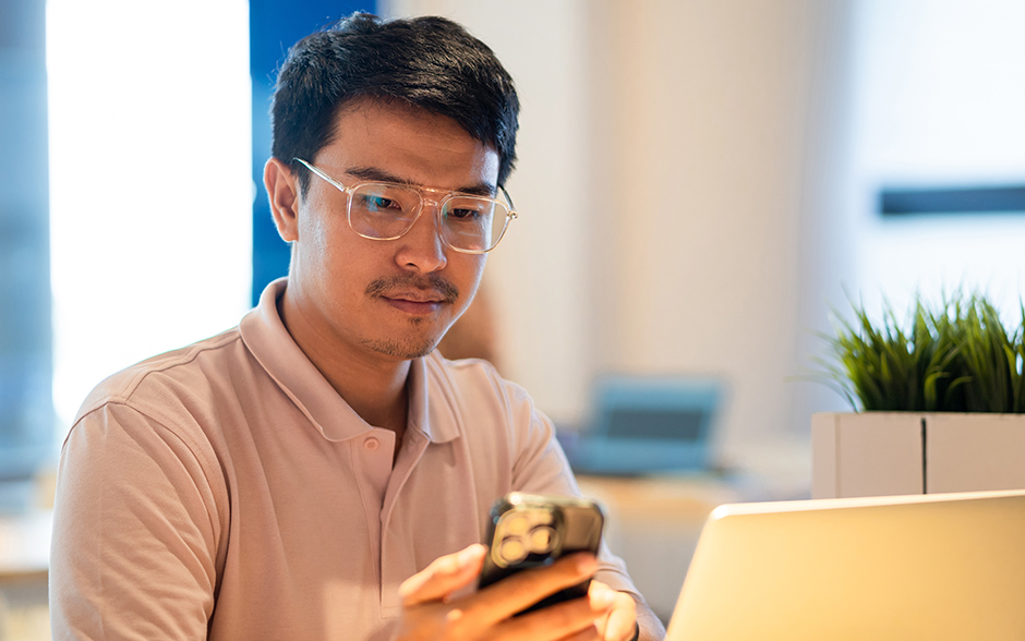 Asian male checks his mobile phone with laptop open.