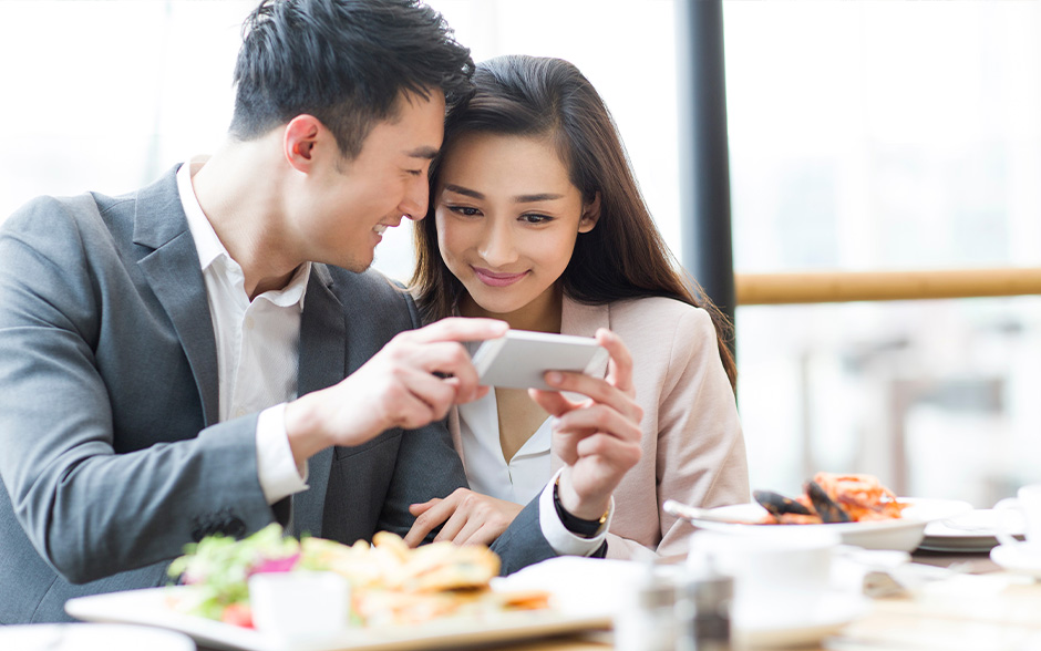 Young Asian couple looking at their smartphone in a restaurant, looking through the perks of a Platinum plan.