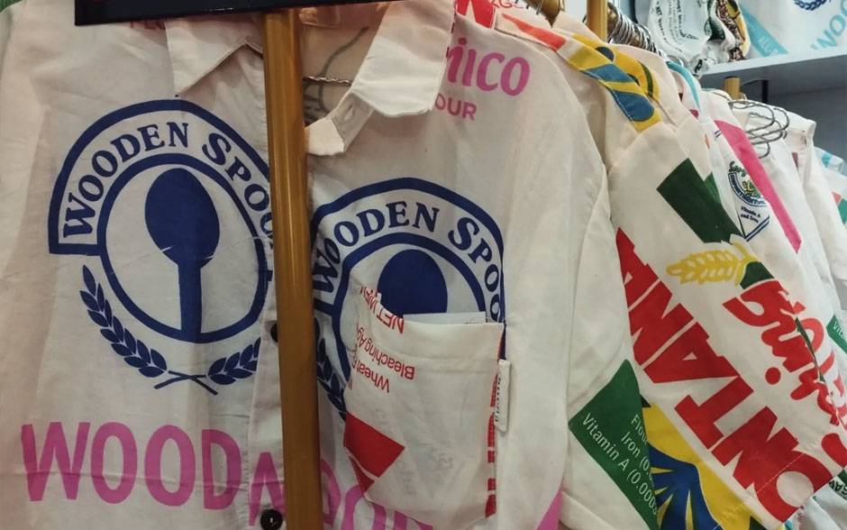 Upcycled clothing pieces from Craftcha, including polo shirts and dresses made from repurposed flour and rice sacks, displayed on a rack.