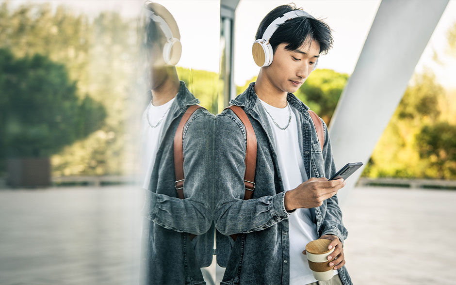 A young man wearing headphones standing outdoors, watching something on his smartphone.