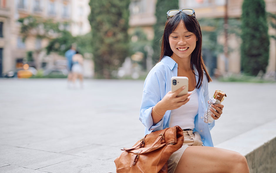 A young woman smiling at her smartphone while having a snack outdoors.