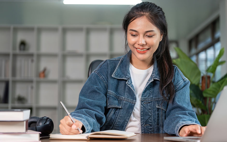 A young Asian woman studies online at home with a laptop and notebook.