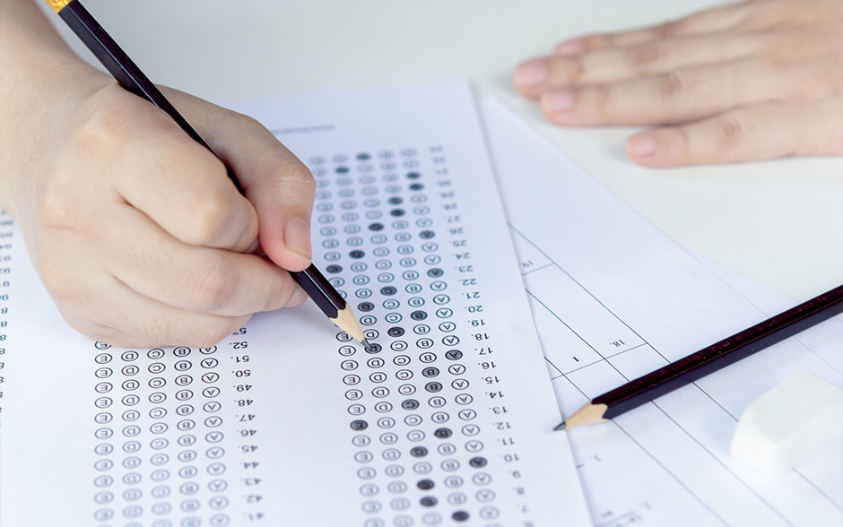 Close-up photo of a hand holding a pencil, shading selected choice on answer sheets.