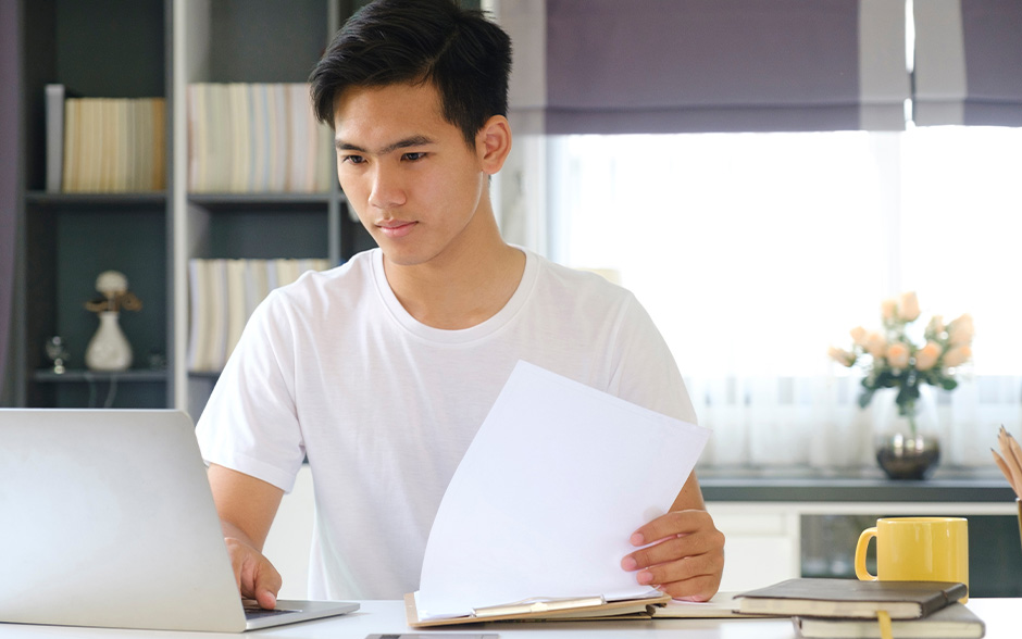 Young Filipino man looking through a clipboard and his laptop at home.