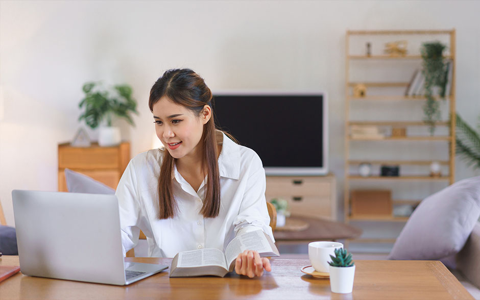 A young Asian woman reading a book and working on a laptop at a modern home office.