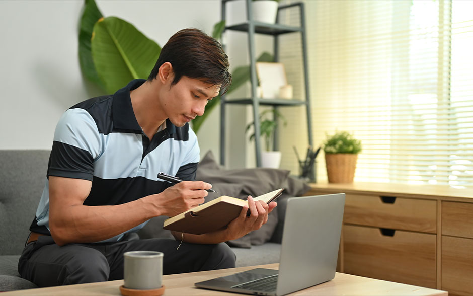 A young Filipino man on the sofa is listing down his tasks in a brown notebook with a laptop in front of him.