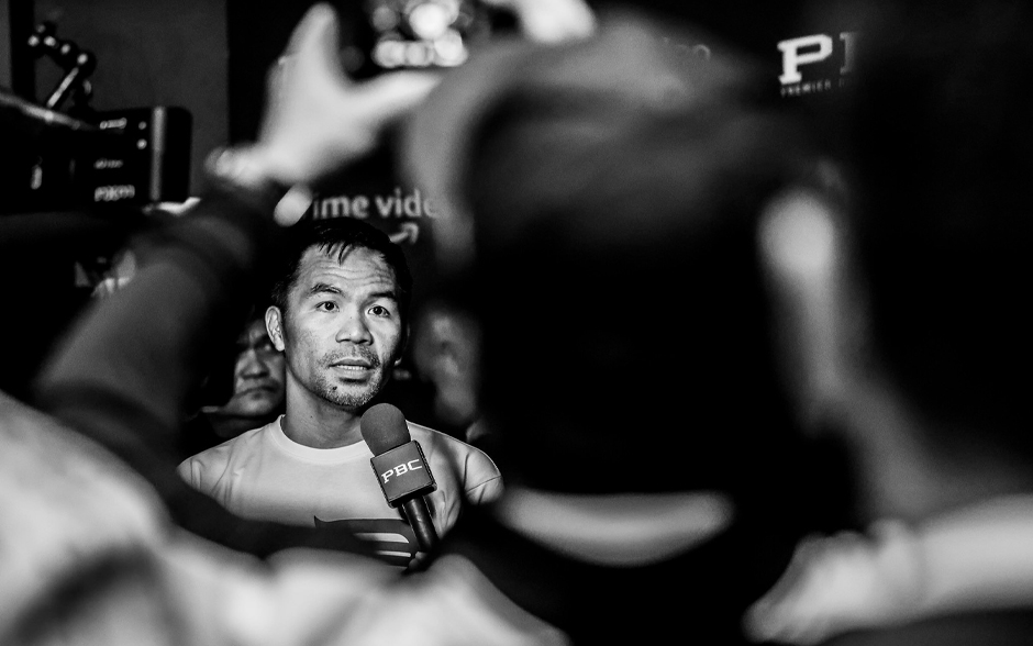  A person speaking into a microphone labeled "PBC" in a crowded setting, with blurred figures in the foreground.