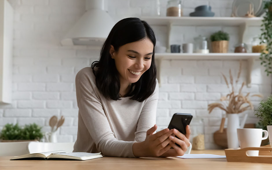Smiling young woman holding a smartphone while sitting at a table, enjoying digital rewards and perks.