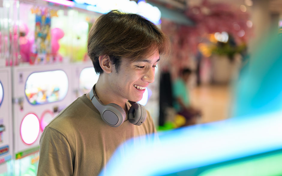 Young man smiling while playing an arcade game, wearing headphones around his neck.