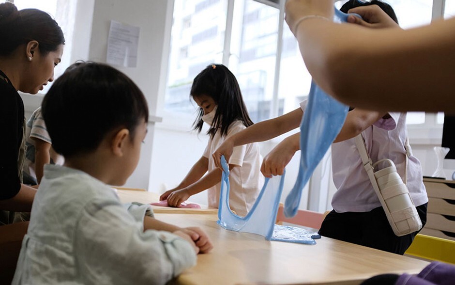 A woman and two children are happily playing with a blue cloth, illuminated by warm, natural light in a cheerful environment.