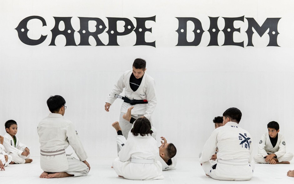 Kids sitting in a Brazilian Jiu-Jitsu class watching their instructor demonstrate their techniques.