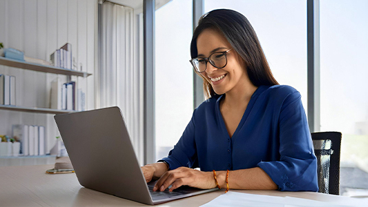 A smiling woman wearing glasses works on a laptop in an office.