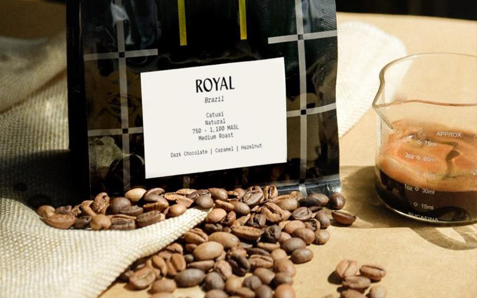 Product photo of a bag of coffee on a table with coffee beans scattered around it and a beaker with a shot of espresso.