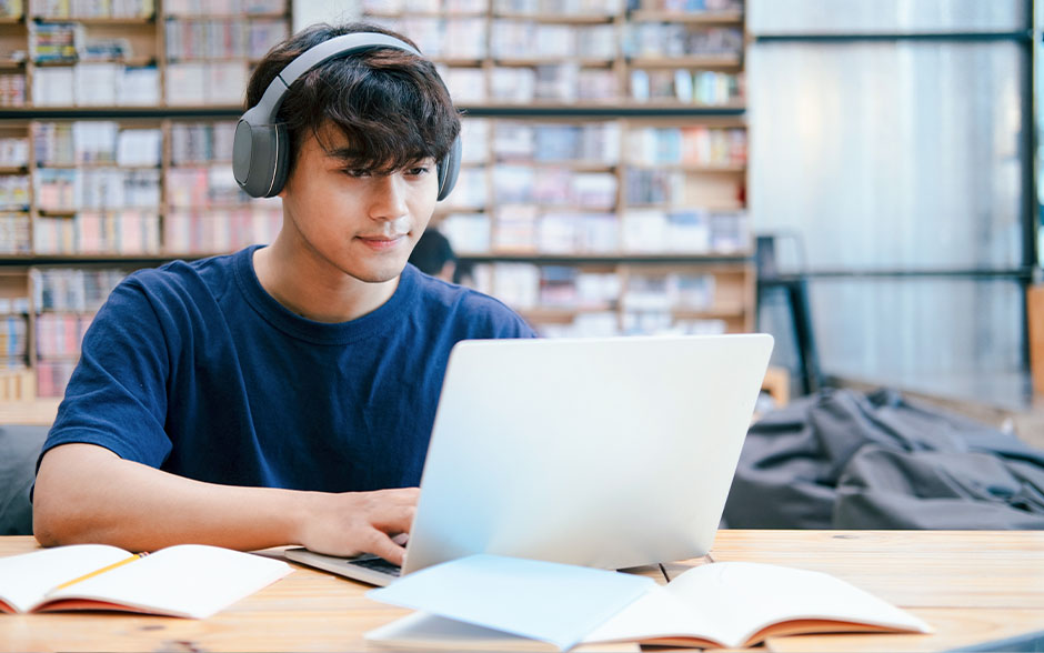 Young student wearing headphones studies on a laptop in a library with open books on the table.