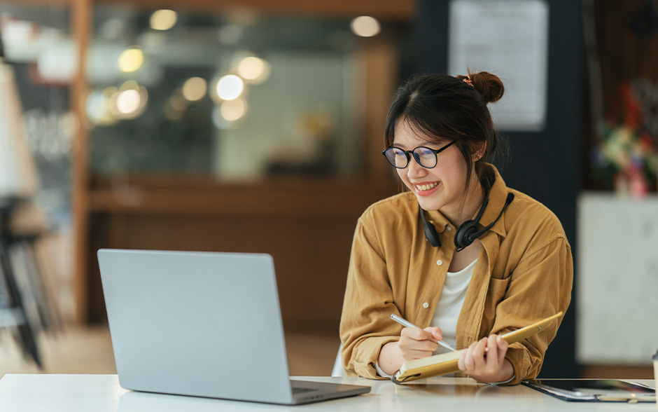 Young woman with glasses taking notes while looking at a laptop