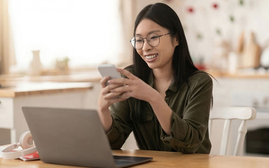Young Asian woman holding her smartphone while using the computer at home.