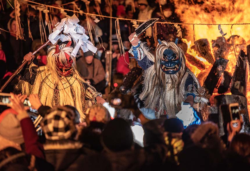 Performers wearing traditional masks and costumes dancing before a bonfire.