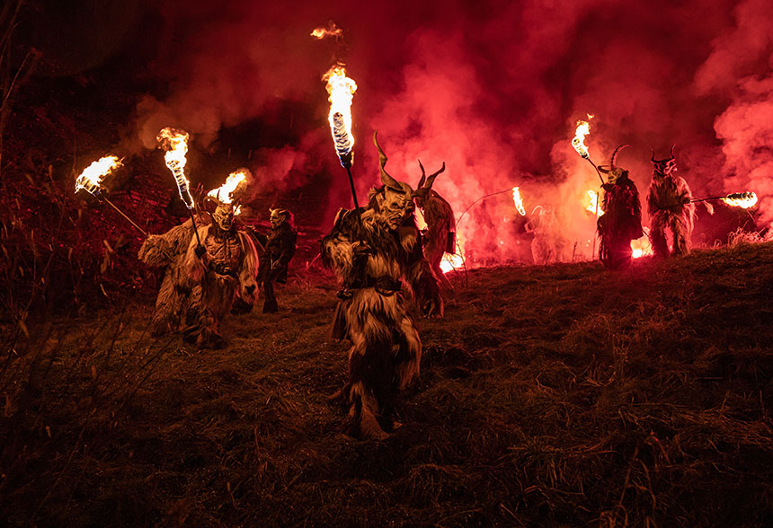 Costumed performers wearing horned masks and fur outfits carry flaming torches through red smoke.
