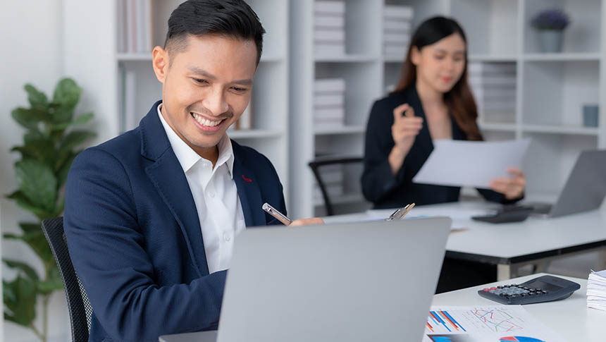 A male professional working on his laptop while a female coworker is behind him.