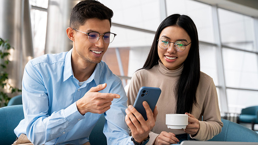 Two colleagues are looking at a smartphone together while sitting on a couch in an office lounge.