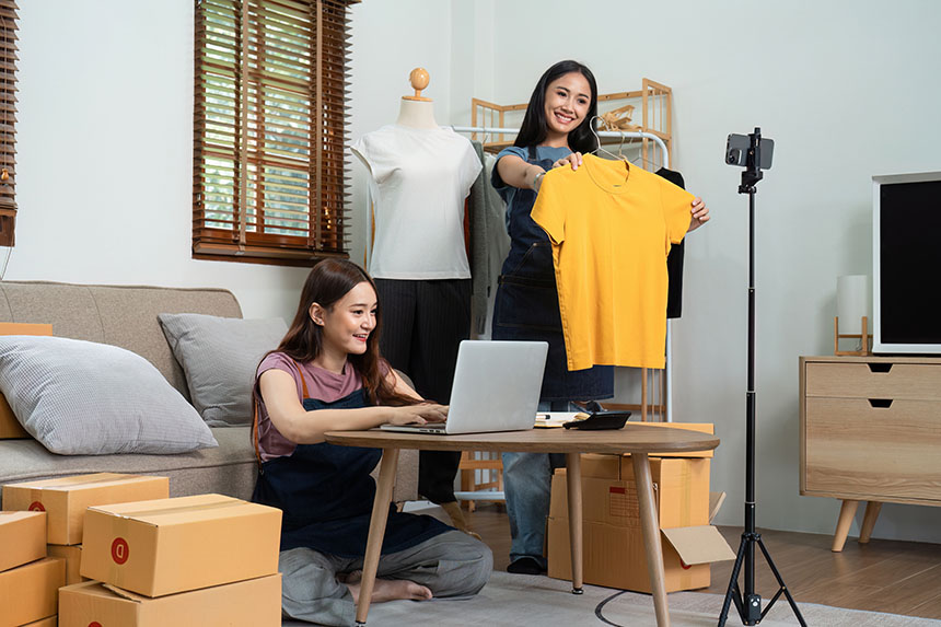 Two girls selling clothes through a live selling platform with their phone and laptop.