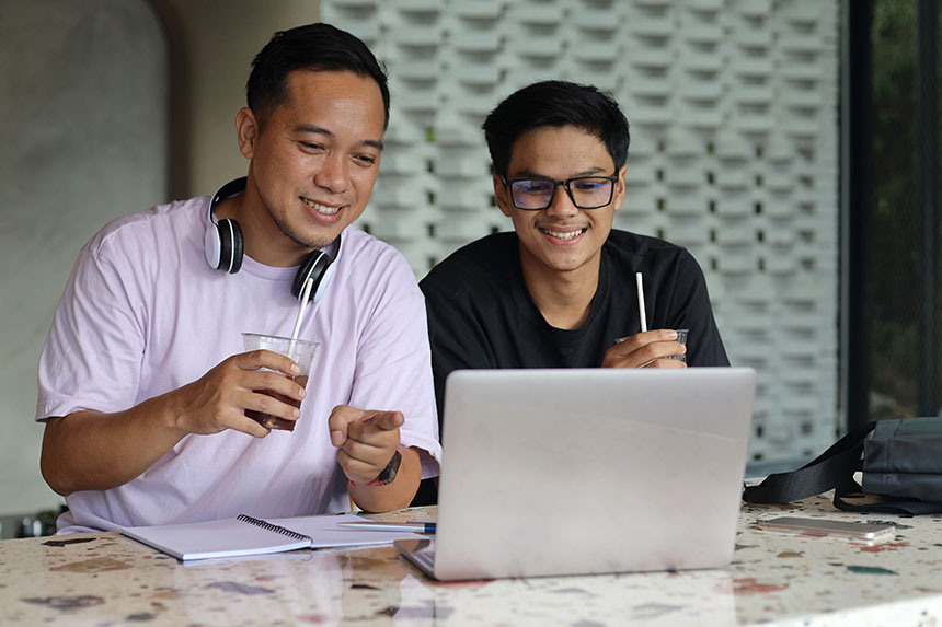 Two men sitting outside a house, using a laptop.
