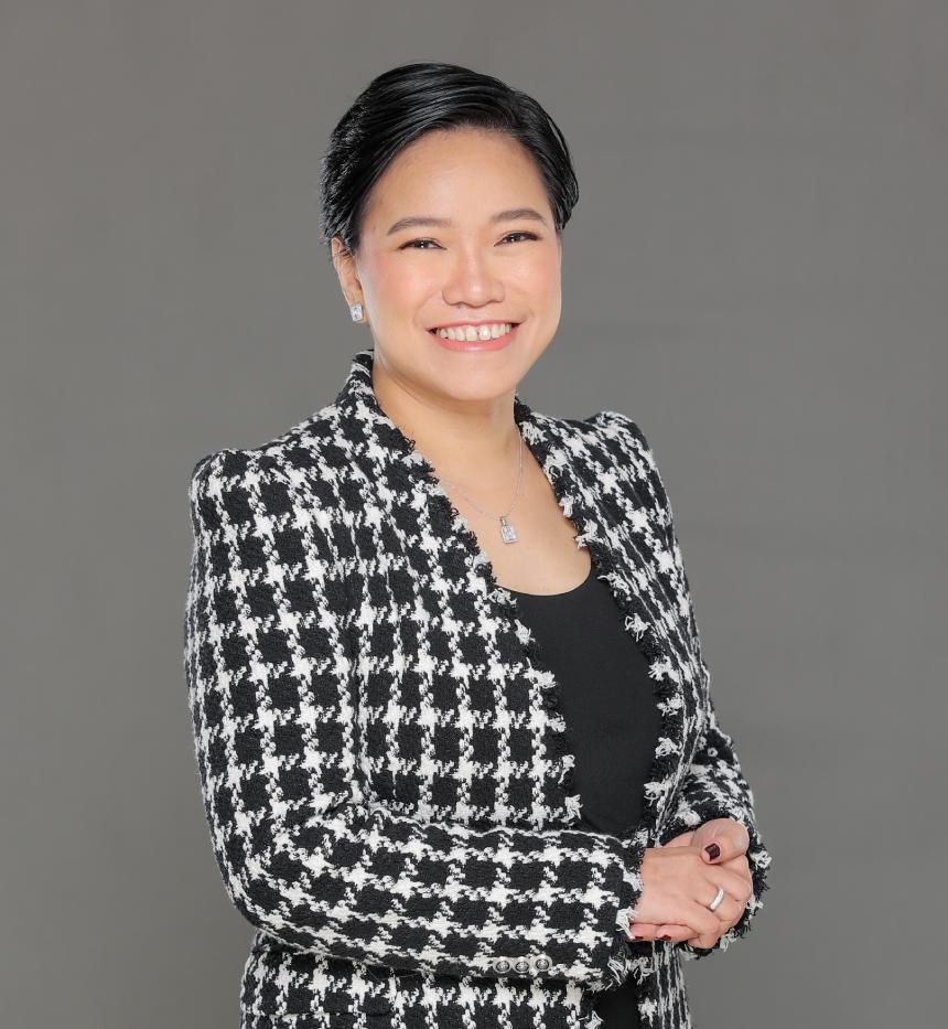 A woman smiling with her hands in her pockets, wearing a plaid blouse against a wall.