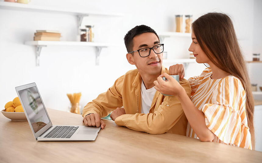 Young Asian couple looking at one another with an open laptop on the table in front of them.