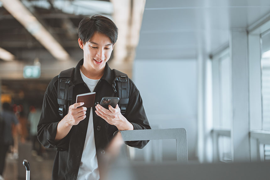 A young man wearing a backpack, smiling and holding his phone, passport, and boarding pass in an airport.