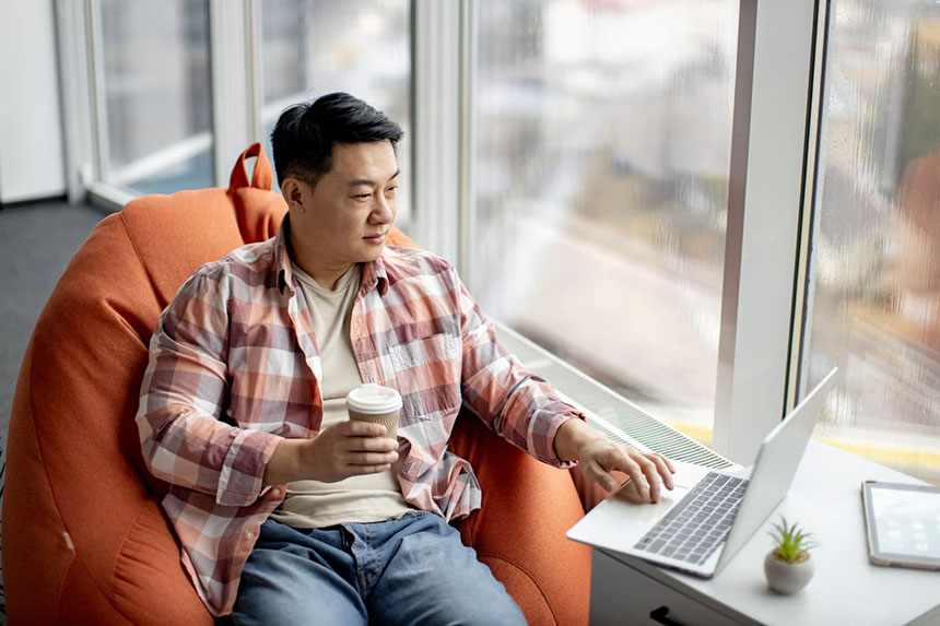 Asian man researching investment options on laptop while looking outdoors.