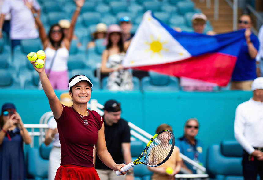 Alex Eala in a maroon kit raising three tennis balls with a smile, the Philippine flag waved behind .
