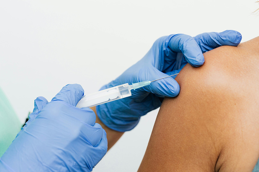 Medical professional wearing gloves injecting a vaccine into the tanned upper arm of a person.