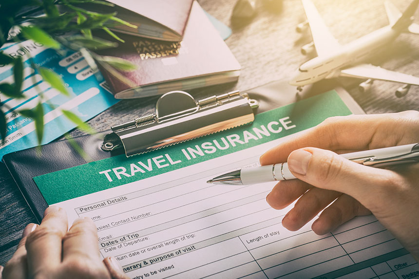 Hand holding pen hovering above a travel insurance form with a passport, boarding pass, and toy airplane on the tabletop.