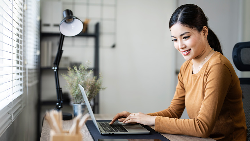 A smiling woman using her laptop while inside her bedroom.
