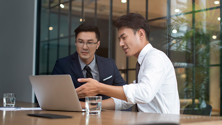 Two professional men talking to each other while using a laptop at an office.