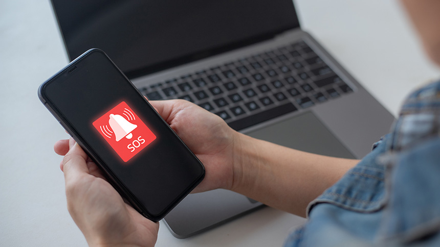 A red alert notification displays at the center of the mobile phone held in front of a laptop.