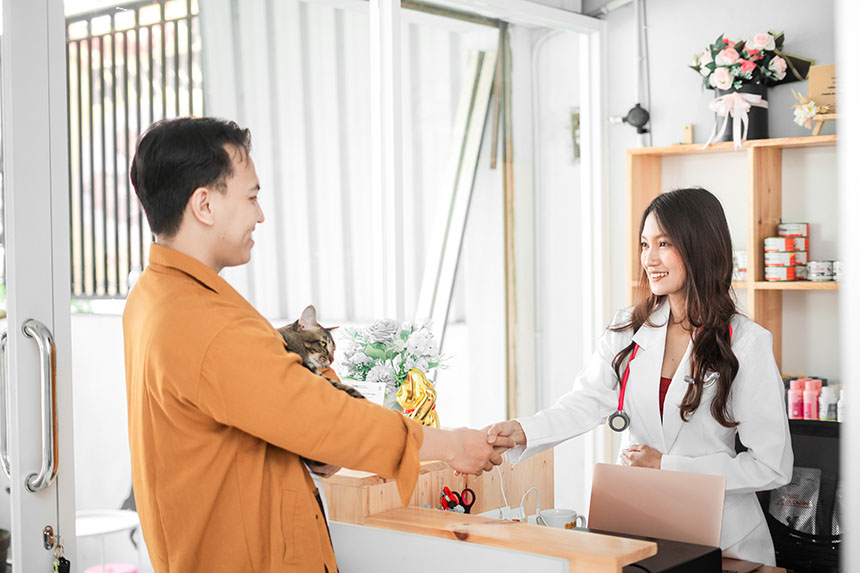 A pet owner holding a cat shakes hands with a veterinarian at a clinic counter.