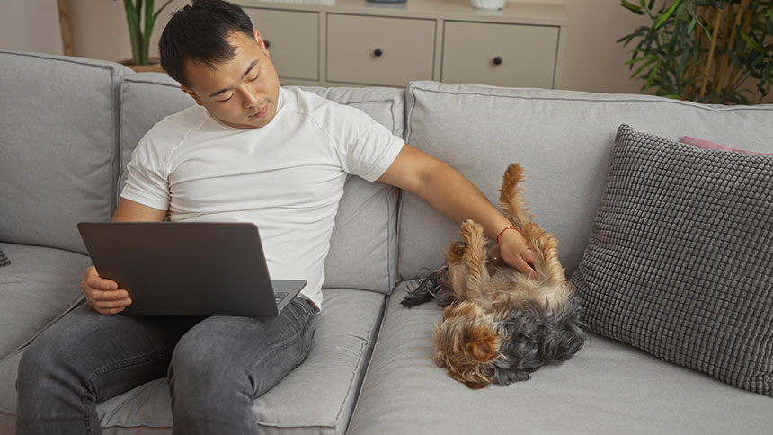 A pet owner relaxes at home with a laptop while bonding with his dog on the couch.