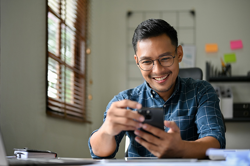 Young Asian man looking at his smartphone while seated on his desk, considering frequently asked questions about his Globe prepaid eSIM.